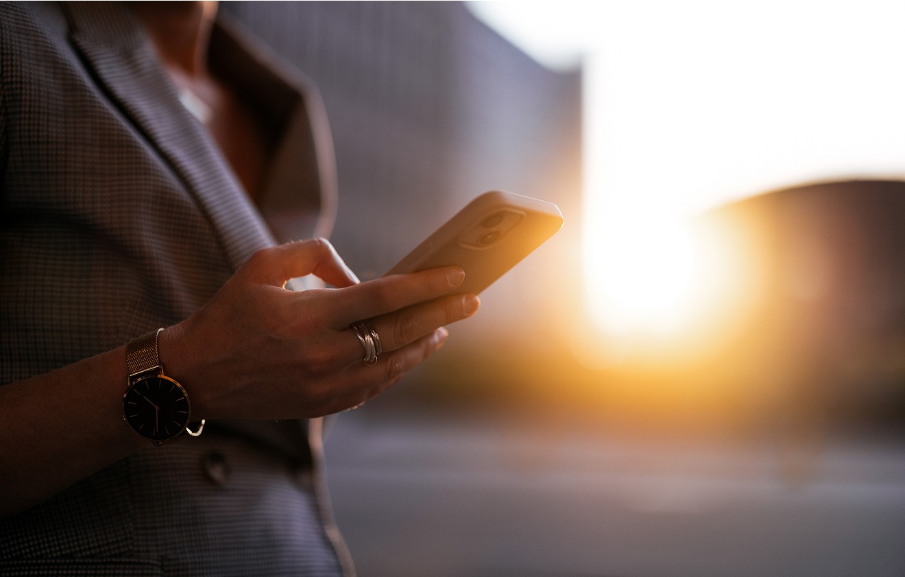 Person wearing a suit and wristwatch holds a mobile phone outdoors at sunset.