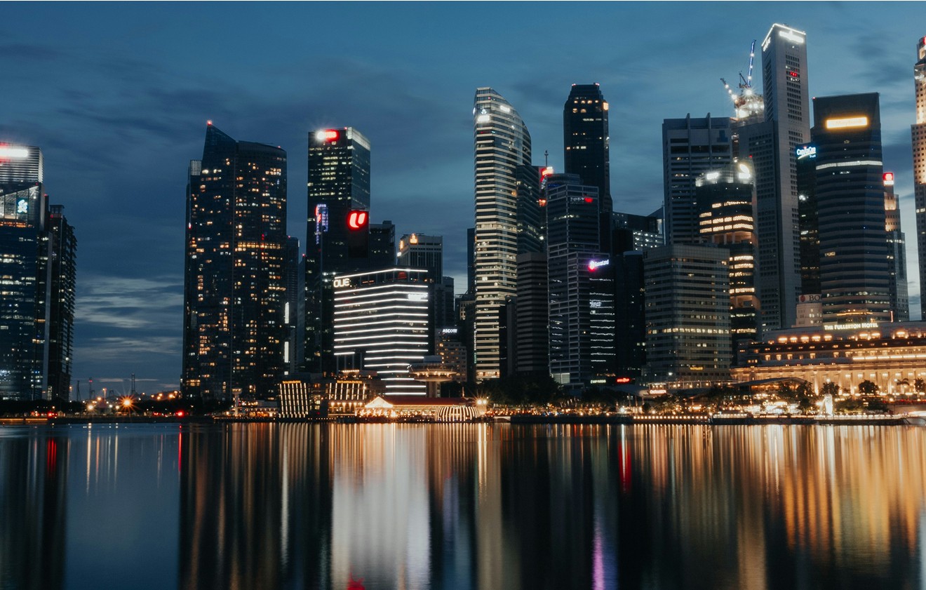 Night-time cityscape of modern skyscrapers with illuminated windows reflected in the calm water below, under a dark blue sky.