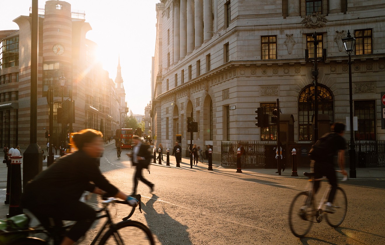 Cyclists and pedestrians move through a city junction at sunset, surrounded by historic and modern buildings.