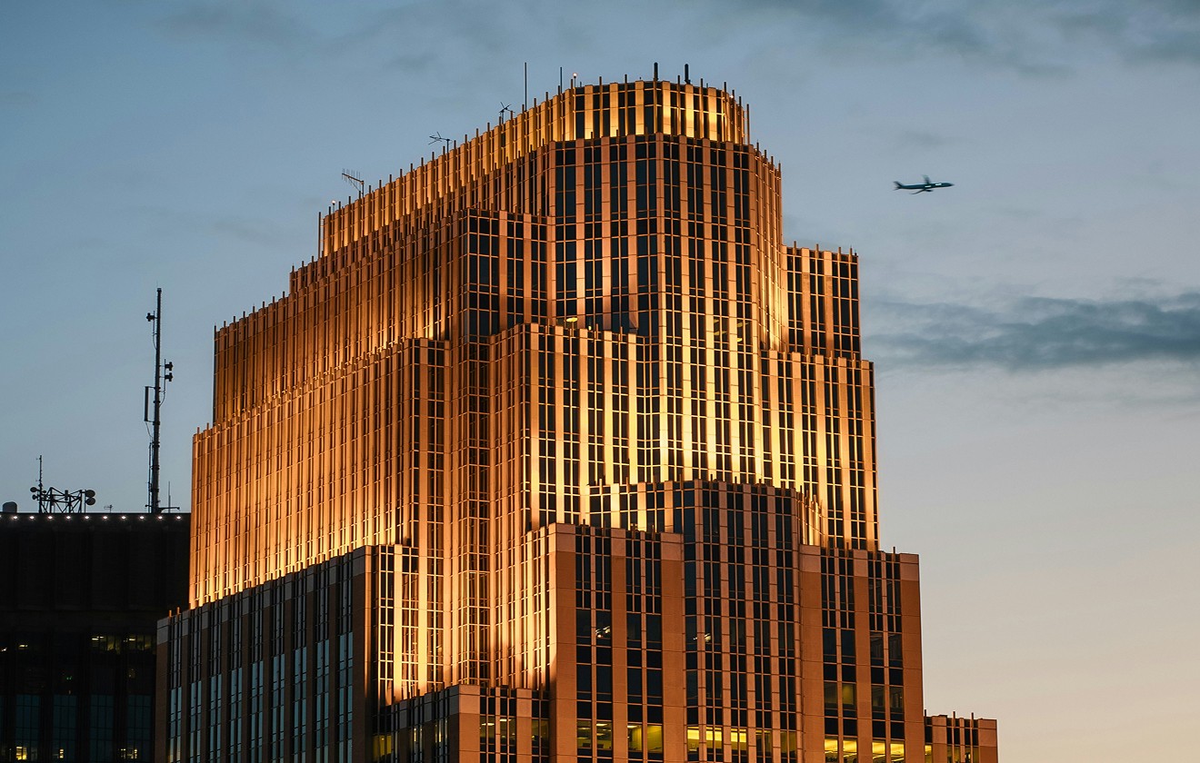 A modern skyscraper is illuminated by warm lights at dusk, with an aeroplane flying in the sky above the building.