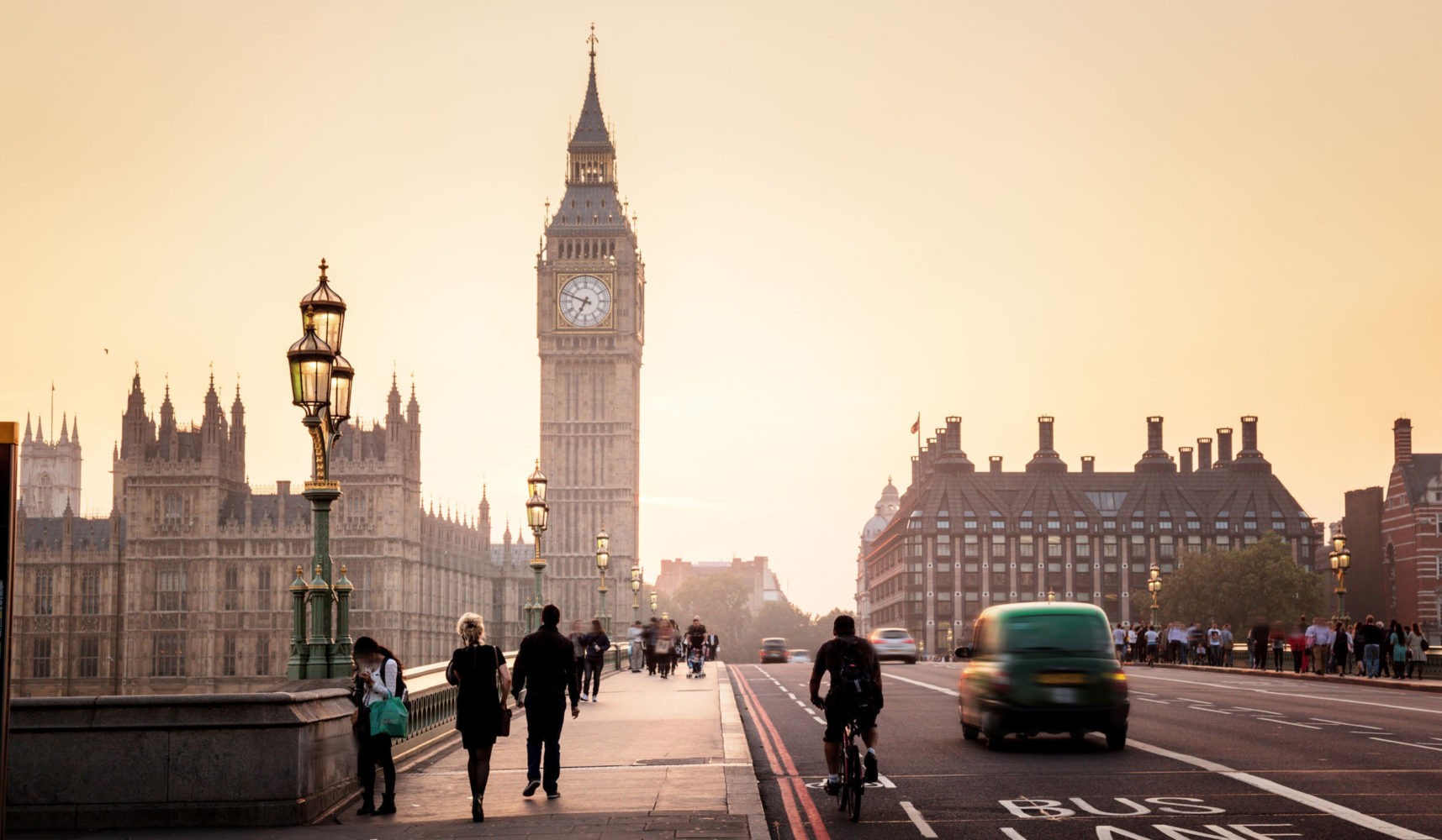 People walk and cycle on Westminster Bridge in London at sunset, with Big Ben and the Houses of Parliament in the background and vehicles on the road.