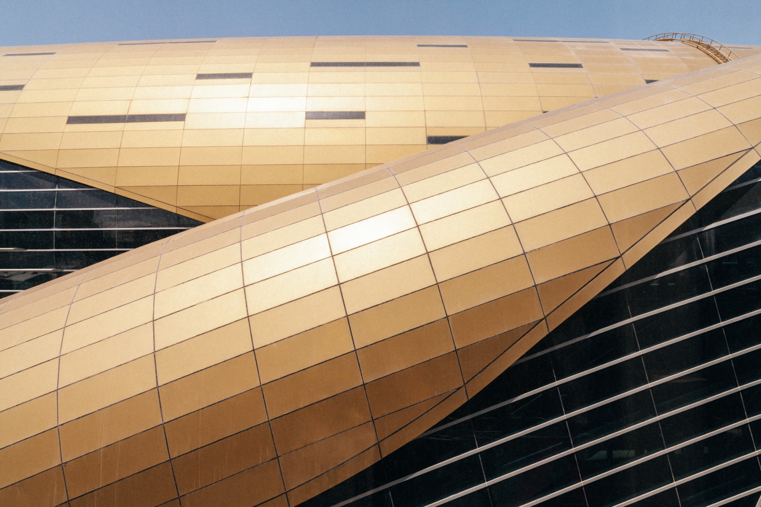 Close-up view of a modern building with overlapping, gold-coloured, curved metal panels and glass sections under a clear sky.