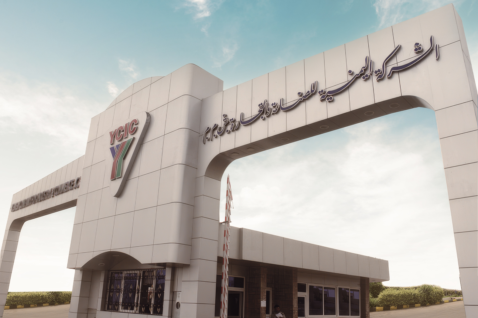 A modern white entrance gate with the logo “YCIC” and Persian text, with a small security kiosk and glass windows underneath, set against a blue sky.